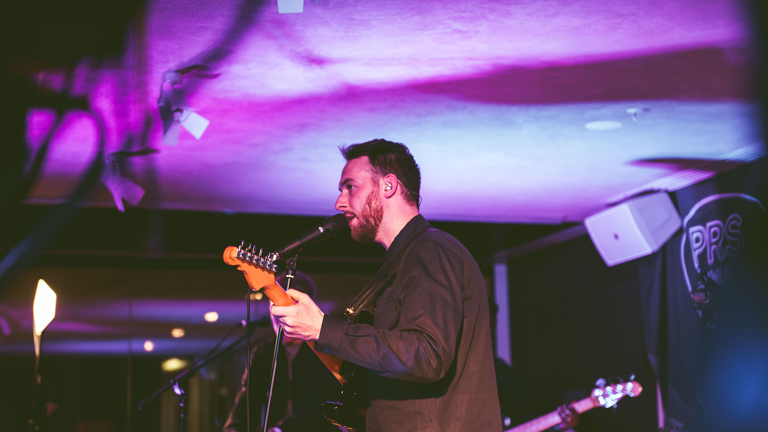 A shot from the side of Honne's Andy Clutterbuck singing and playing guitar on stage at PRS Presents