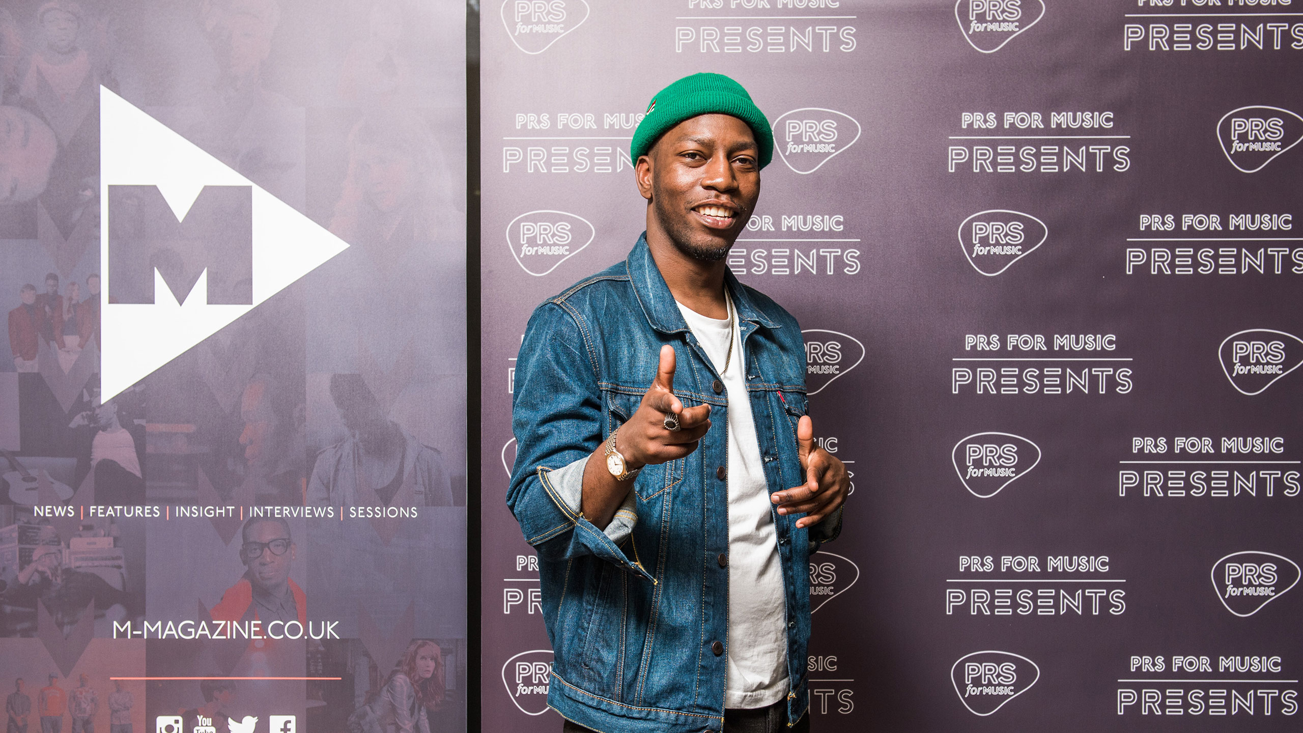 Tiggs Da Author points at the camera in front of the PRS for Music backdrop, wearing a green beanie hat, white t shirt and denim jacket
