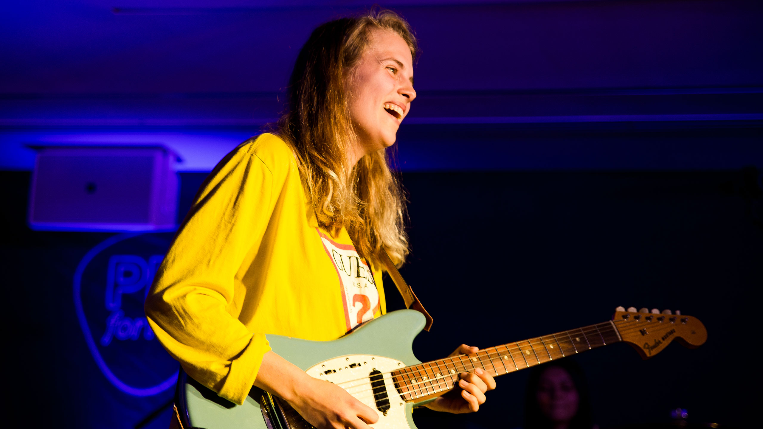 Marika Hackman, wearing a bright yellow shirt and holding her guitar, smiles at the audience on stage at PRS for Music Presents