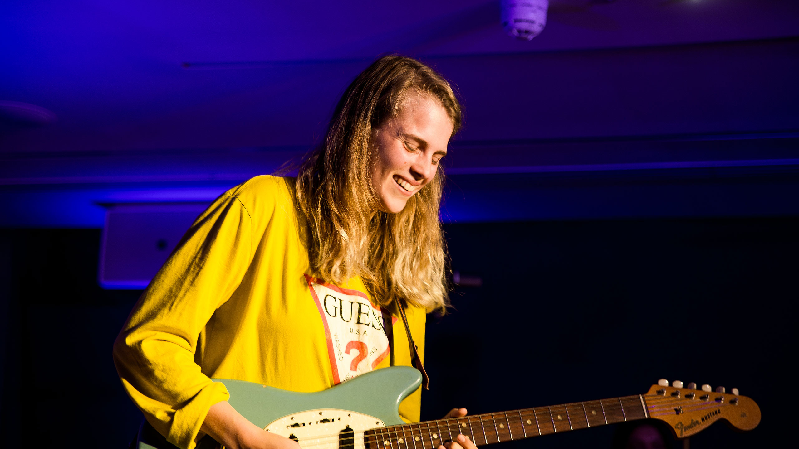 Marika Hackman, wearing a bright yellow shirt and holding her guitar, smiles at the floor on stage at PRS for Music Presents