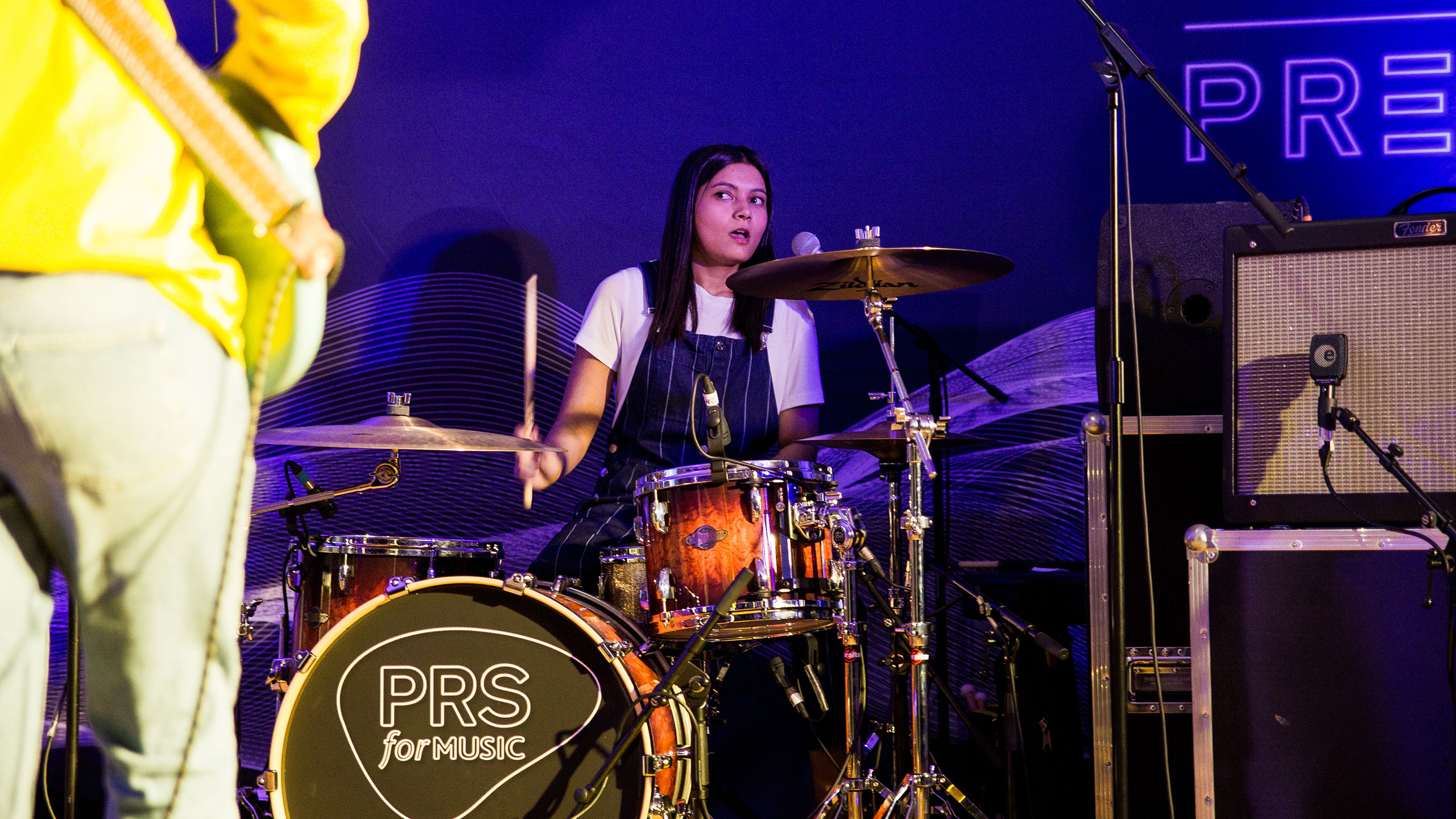 Marika Hackman's drummer, wearing a white t shirt and dungarees, looks up whilst playing live at PRS for Music Presents