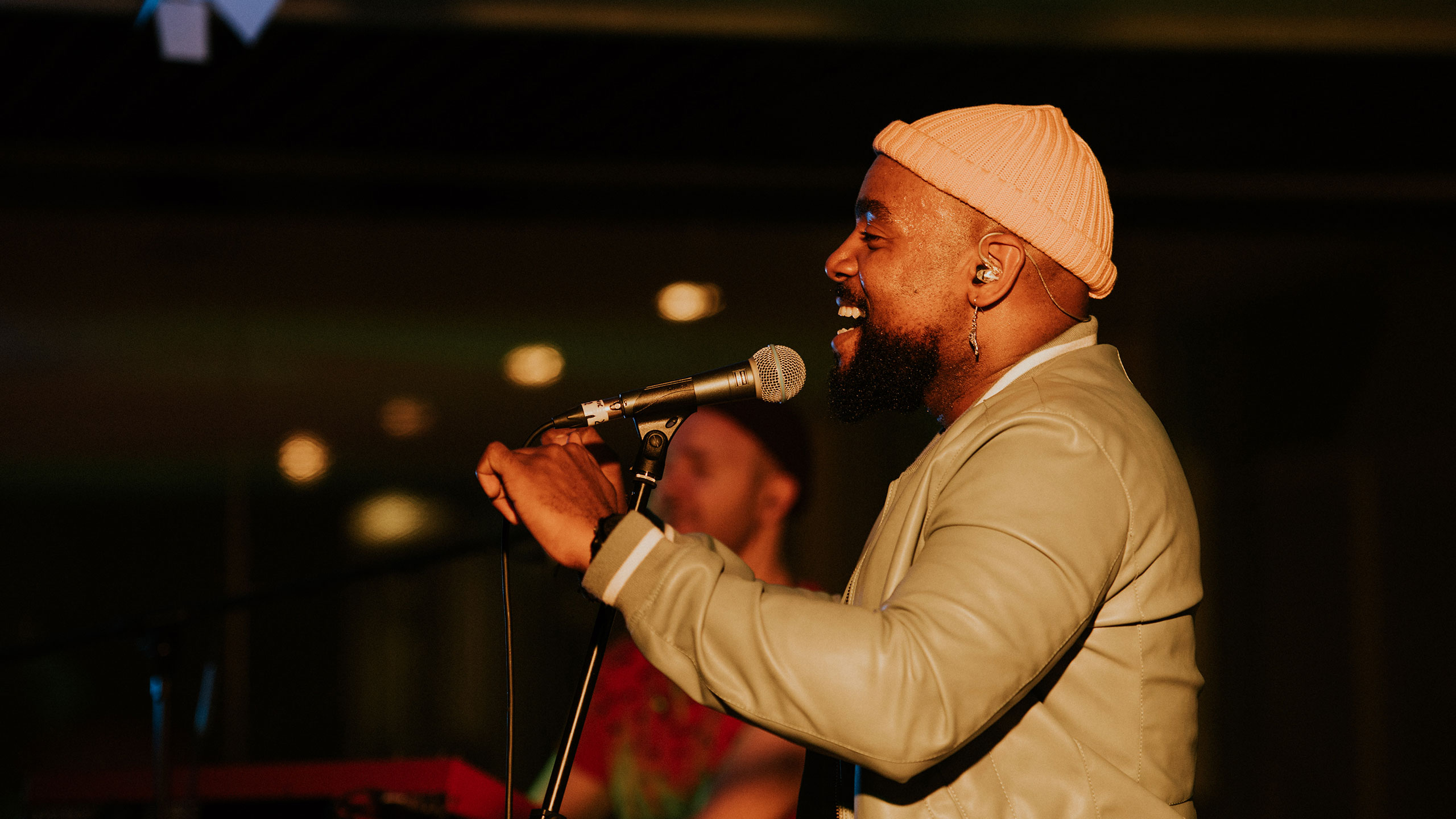 Jodie Abacus, wearing an orange beanie hat and grey jacket, dances and sings into the microphone at PRS for Music Presents
