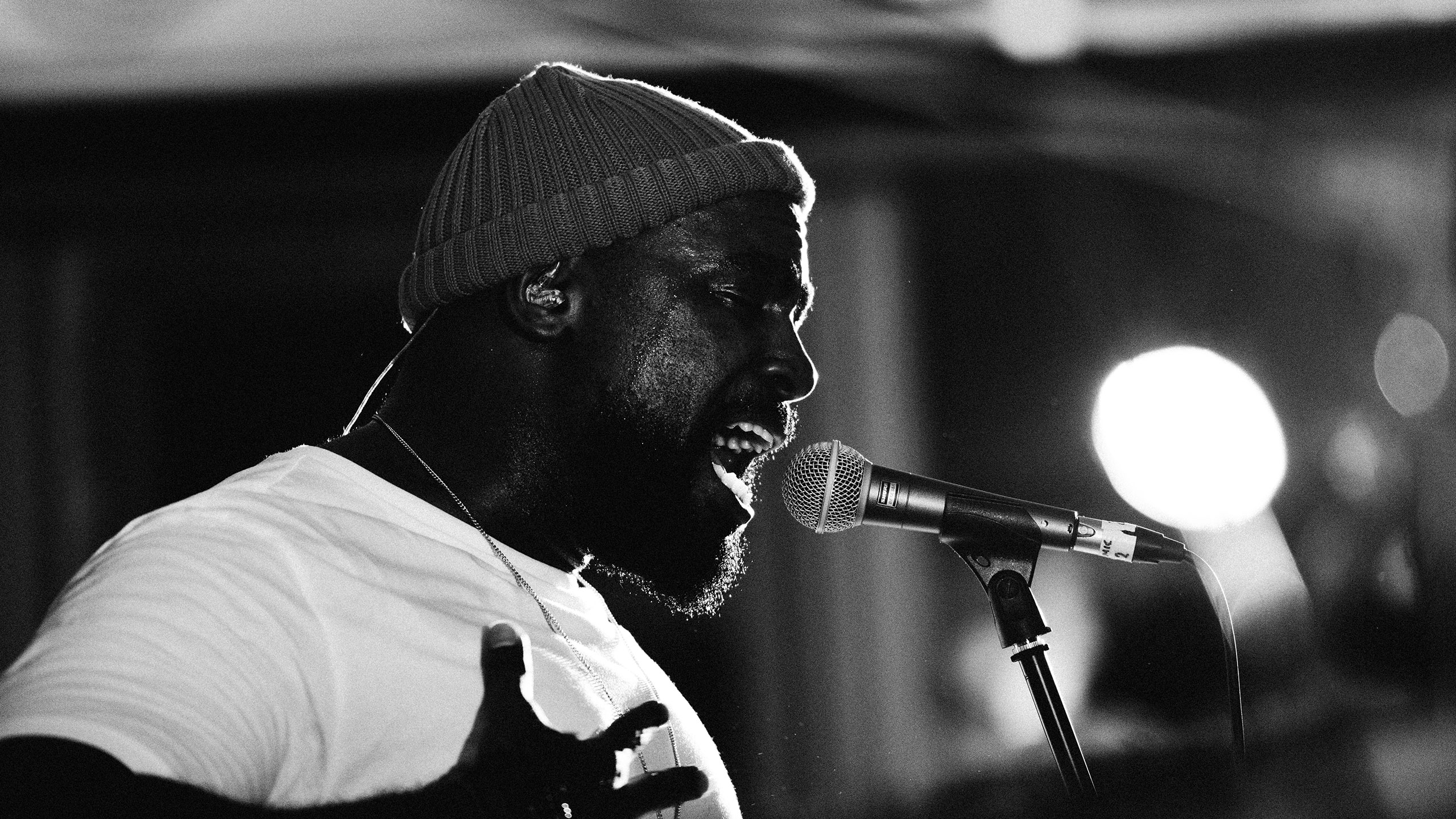 A black and white photo of Jodie Abacus in a white t-shirt and beanie hat singing and stretching his hand at PRS for Music Presents