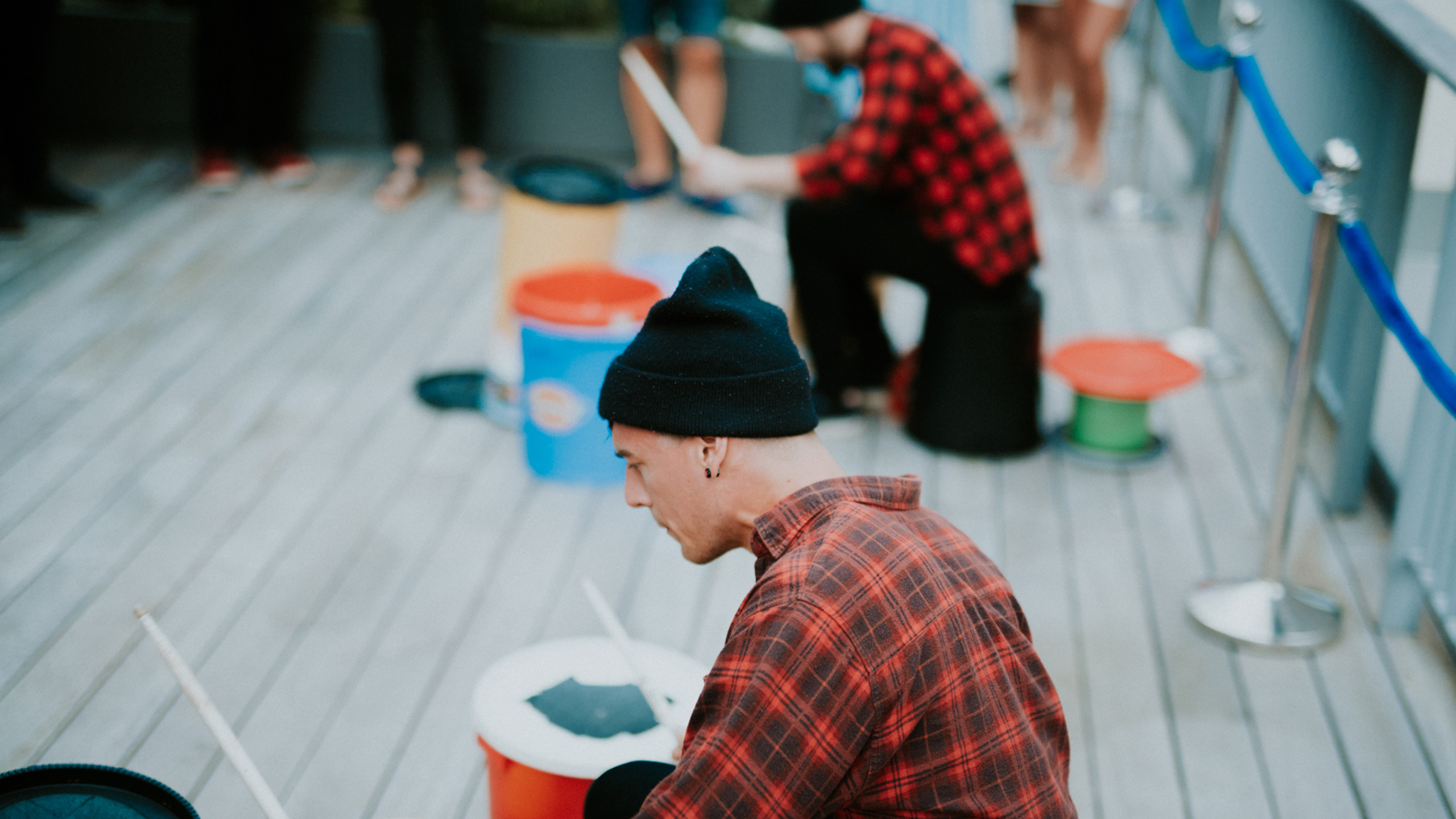 Two men in red checked shirts and black beanies drum on tubs with drum sticks