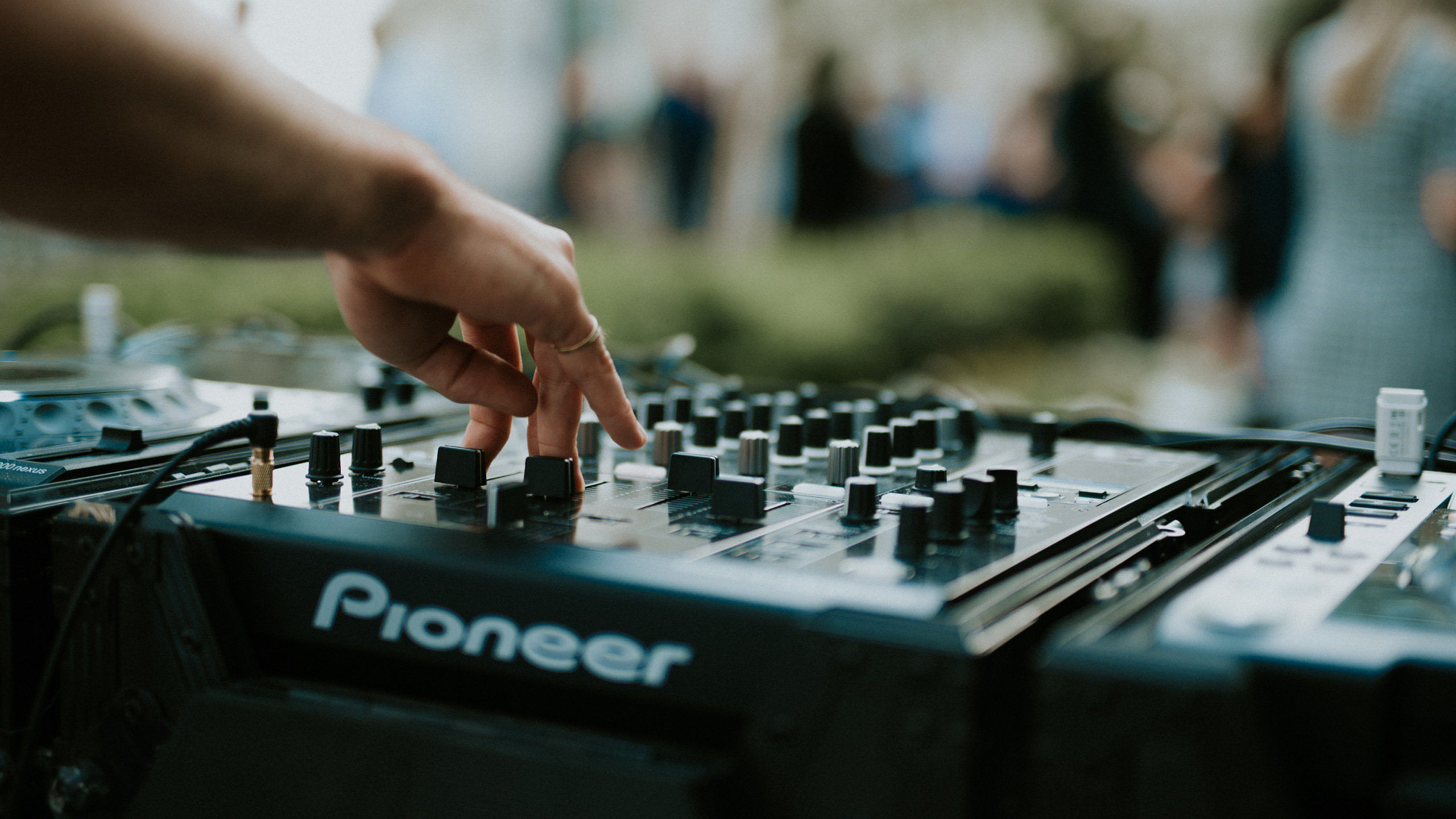 A hand on a mixing desk with people and plants in the background